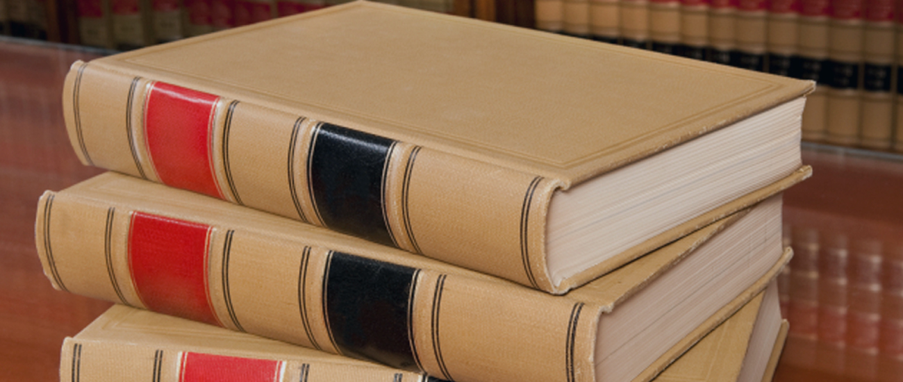 Stack of beige hardcover books with red and black spines on a wooden table, with a bookshelf in the background.